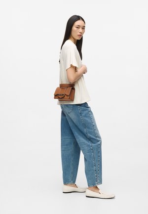 Mujer con camiseta blanca holgada, vaqueros azules de pierna ancha, mocasines blancos y llevando un pequeño bolso cruzado de cuero marrón con cadena dorada.