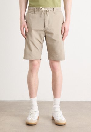 Pantaloni scurți bej dintr-un material texturat, cu o talie elastică dotată cu șnur. Purtați cu adidași albi și șosete albe.