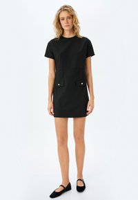 Vestito nero con maniche corte, scollatura tonda, due tasche frontali con bottoni dorati e una silhouette aderente. Indossato con ballerine nere.