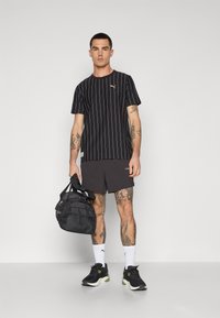 Sort vertikalt stribet t-shirt med logo, grå shorts, sort sportstaske og sorte sneakers med grønne accenter; med tatoveringer påarmene.