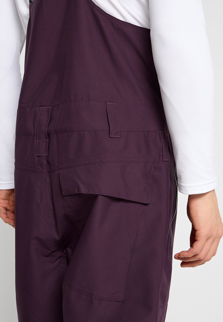 Fioletowe ogrodniczki o gładkiej fakturze, z boczną kieszenią i regulowanymi ramiączkami. Wzór zawiera kontrastową białą koszulę z długim rękawem pod spodem.