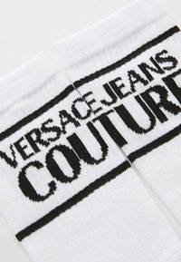 Baltas zeķes ar melnu "Versace Jeans Couture" logo, adīts tuvu augšējai malas līnijai uz vienkrāsaina fona.