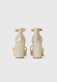 Zlate bleščeče sandale z visokim petom in širšim podplatom, z zaponko ob gležnju in odprtimi prsti. Gladek teksturiran material z odsevnimi površinami in nastavljivim zaponkom.