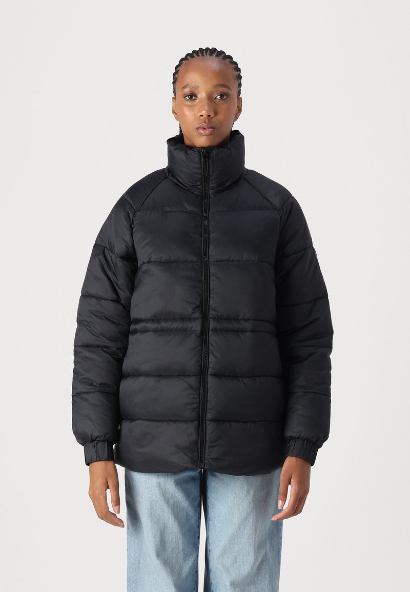 Casaco puffer preto com gola alta, apresentando um fecho zip na parte da frente, design acolchoado e punhos elásticos. Usado sobre uns jeans azul claros.
