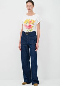 Camicia bianca con stampa floreale colorata, abbinata a pantaloni in denim scuri a vita alta con gamba larga e una cintura con una grande fibbia.