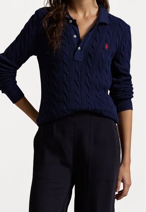 Femme portant un pull en maille torsadée bleu marine avec col et logo rouge, assorti à un pantalon noir ajusté, debout avec une main sur la hanche.