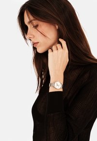 Femme portant un haut en tricot noir touchant ses cheveux, avec une montre-bracelet argentée à cadran rond et bracelet en métal au poignet gauche.