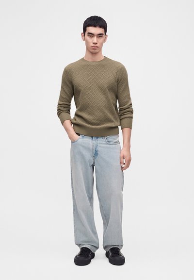 Jeune homme portant un pull olive texturé, un jean bleu clair ample, des baskets noires, debout avec une main dans la poche sur fond blanc.