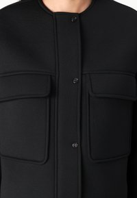 Haine exterioare negre cu guler rotund, două buzunare frontale și închidere cu nasturi. Fabricat dintr-un material texturat cu un design croit.