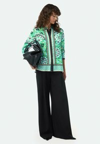 Gemusterte grüne Bluse mit schwarzen Akzenten, kombiniert mit weiten schwarzen Hosen, hält eine schwarze Tasche. Glattes Material und lebhaftes Blumenmuster.