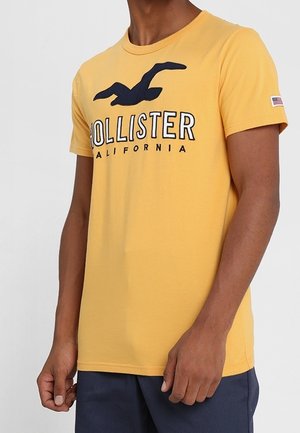Camiseta de algodón amarilla con un logo de "Hollister California" en azul marino y un diseño de gaviota. Incluye un pequeño acento de bandera estadounidense en la manga.