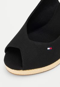 Scarpa nera in tela con punta aperta e suola in gomma testurizzata. Presenta un piccolo logo ricamato in rosso, bianco e blu.
