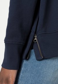 Marinblå sweatshirt med ribbade fållar och muddar, med en sidDragkedja i metall. Texturerad tyg, ihop med denim jeans.