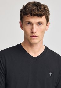 Camiseta negra de cuello en V, hecha de algodón suave, con un pequeño detalle bordado de una figura en el lado izquierdo del pecho. Diseño simple y moderno.