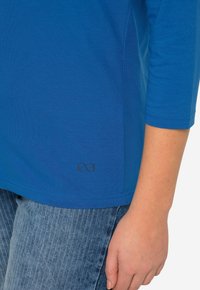 Modré tričko s dlouhým rukávem vyrobené z měkké látky s kulatým výstřihem. Na lemu má decentní logo nekonečna a je doplněno světle modrými džíny.