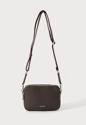 Μικρή τσάντα crossbody από σκούρο καφέ δέρμα με ρυθμιζόμενο λουρί και ασημί φερμουάρ, λογότυπο Calvin Klein μπροστά.