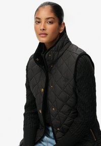 Czarna pikowana kamizelka z złotymi guzikami, wysokim kołnierzem i teksturowanymi czarnymi rękawami swetra. Noszona na dżinsach dla uzyskania warstwowego wyglądu.