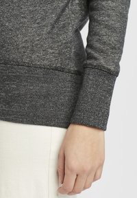 Mørkegrå hettegenser med et teksturert, flekkete mønster. Har ribbestrikkede mansjetter og kant, og viser en uformell, tettsittende design.