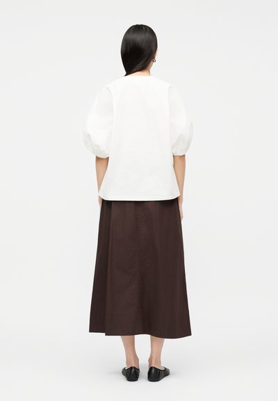 Γυναίκα με μακριά μαύρα μαλλιά που φοράει λευκό μπλουζάκι με φουσκωτά μανίκια, καφέ φούστα μέση και μαύρα παπούτσια με επίπεδη σόλα, στέκεται γυρισμένη με την πλάτη σε ένα απλό φόντο.