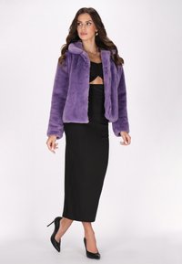 Giacca in finta pelliccia viola con collo accogliente, maniche lunghe e vestibilità rilassata. Indossata sopra un vestito nero aderente e tacchi alti neri.