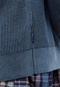 Niebieski sweter dziany z teksturowanym wzorem, ściągaczami u dołu, bocznym rozcięciem i małą metką marki. Połączony z wytłaczanymi spodniami w kratę.