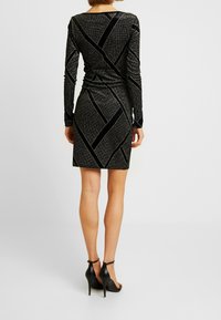 Schwarzes Langarmkleid mit figurbetontem Schnitt, aus einem strukturierten, glitzernden Stoff mit kontrastierenden, diagonalen Samtstreifen. Absatzsandalen.