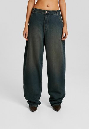 Kobieta ubrana w wysokie, luźne dżinsy w ciemnym niebieskim kolorze z wyblakłymi detalami, w połączeniu z brązowymi spiczastymi butami, na jednolitym tle.