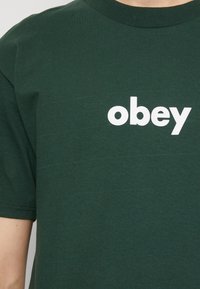 T-shirt en coton vert avec encolure ronde ; texte blanc "obey" imprimé sur le devant ; texture lisse et manches courtes.