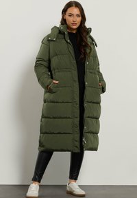 Grøn pufferjakke med hætte, der har et langt design, knap- og lynlåslukning samt sidelommer. Båret over sorte leggings og sneakers.