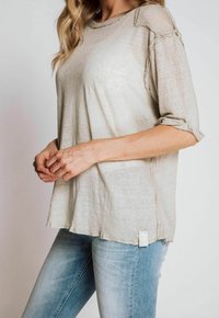 Beige Kurzarmoberteil in lockerer Passform aus leichtem Stoff, mit rohen Kanten und subtiler Textur, kombiniert mit hellblauen Jeans.