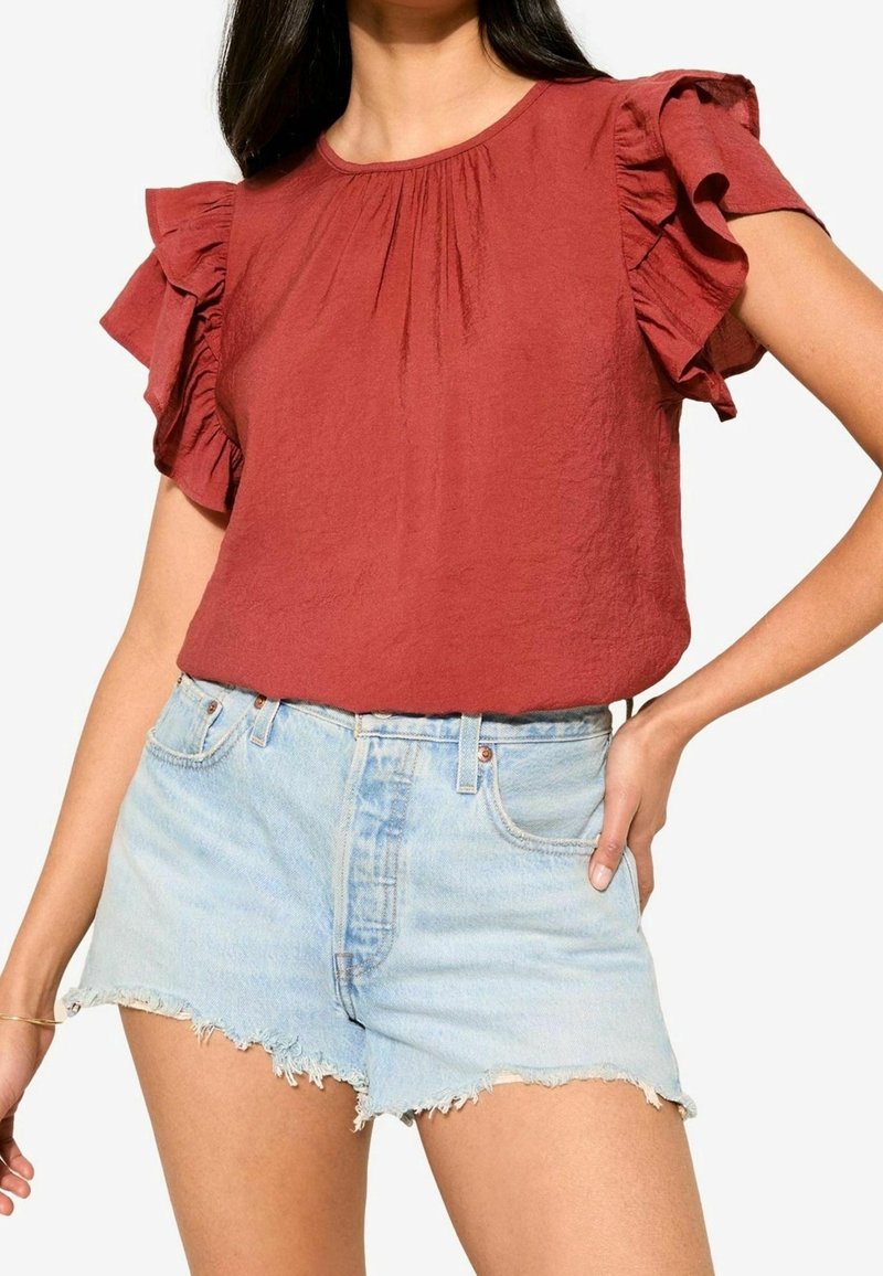 Femme portant un chemisier à manches volantées de couleur rouille, rentré dans un short en jean bleu clair taille haute avec des ourlets effilochés.