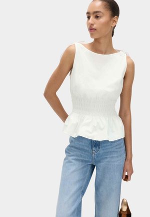 Kvinde iført en ærmeløs hvid peplumtop med markeret talje og lyseblå jeans, står med hånden i lommen mod en ensfarvet baggrund.