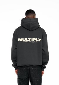 Sudadera negra oversized con capucha ajustable, con gran texto en crema "MULTIPLY" en la parte posterior. Combinada con jeans de mezclilla oscuros.