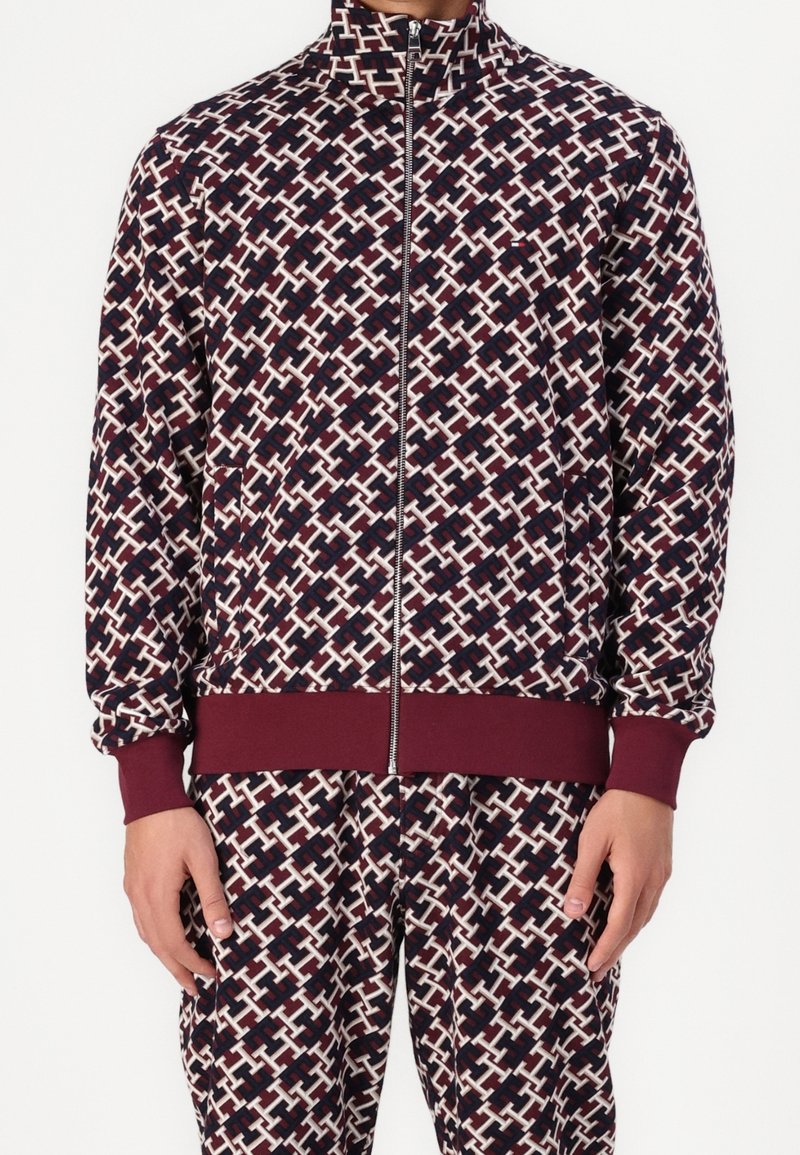 Chaqueta de poliéster con cremallera, con un patrón geométrico en azul marino, blanco y burdeos. Presenta puños y dobladillo acanalados, y bolsillos laterales.