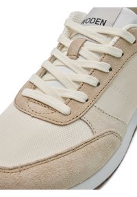 Nahaufnahme eines beigen und cremefarbenen Sneakers mit strukturiertem Mesh-Vorderteil, Wildledereinsätzen, weißen Schnürsenkeln und einem "WODEN"-Label auf der gepolsterten Zunge.