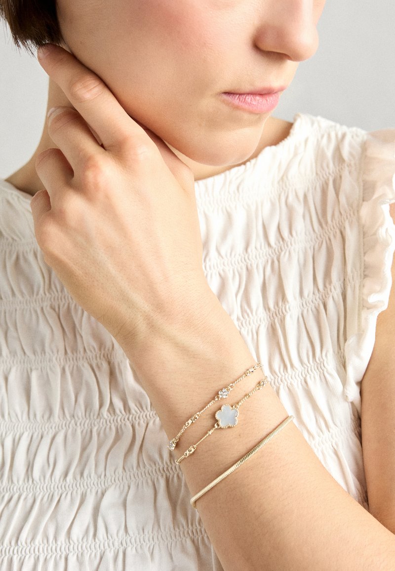 Gold-tone bracelets on a wrist, featuring a flower-shaped charm with a light blue stone and small, clear accents. Textured white blouse background.