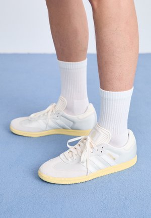 Jambes portant des chaussettes blanches mi-mollet et des baskets blanches avec des semelles jaunes, posées sur un sol recouvert d'un tapis bleu clair.