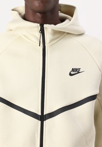 Haut à zip Nike en tissu crème avec une fermeture éclair noire et une bande noire horizontale. Comprend une capuche avec cordon de serrage et un logo de la marque sur la poitrine.