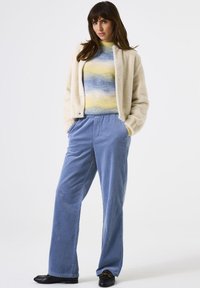 Eine Frau trägt eine cremefarbene Fleecejacke über einem gewellten blau-gelben Pullover, weit geschnittene blaue Cordhosen und schwarze Slipper und steht mit den Händen in den Taschen.