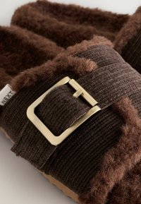 Chaussons en tissu marron avec des accents doux et duveteux, une texture côtelée et un détail de boucle métallique. Le design est confortable avec une doublure intérieure beige.