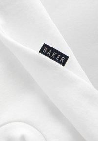Fehér pamut anyag, látható redővel. Egy sötétkék címke mutatja a "BAKER" feliratot fehér betűkkel, jelezve a termék márkáját.