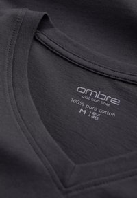 Šedé tričko s výstřihem do V a hladkým povrchem, vyrobené ze 100 % bavlny. Na štítku je nápis "ombre bavlněná řada", velikost M, s uvedenými pokyny k péči.