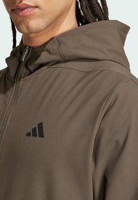 Brun zip-up hoodie med høj krave, lavet af glat stof. Har et sort trekantet logo på brystet og diskrete syninger.