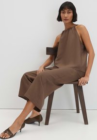 Woman with short dark hair sitting on dark wooden chair wearing sleeveless brown top, matching wide-legged pants, and brown heeled sandals.