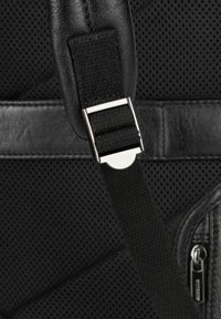 Cinghia nera dello zaino con un pannello in nylon testurizzato e una fibbia metallica lucida, con una finitura in pelle liscia nei dintorni. Dettaglio della cerniera visibile.