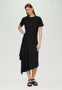 Zwarte midi-jurk met een ronde hals, korte mouwen, asymmetrische zoom en gedrapeerde details aan één kant. Draag het met zwarte platformsandalen.