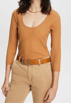 Femme portant un haut à manches longues côtelé, couleur rouille, rentré dans un pantalon beige avec une ceinture en cuir marron et un collier chaîne doré.