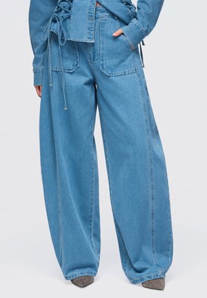 Kvinde iført lyseblå denim bukser med vide ben og store forlommer, kombineret med grå spidse sko, stående mod en ensfarvet baggrund.