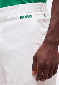 Το δεξί χέρι ενός σκουρόχρωμου ατόμου ξεκουράζεται κοντά στην τσέπη λευκού παντελονιού με πράσινη κεντητική "BORG", φορώντας πράσινο πουκάμισο.