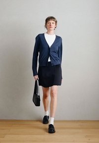 Marineblauwe cardigan over een wit topje, zwarte rok, zwarte schoenen en witte sokken. Draagt een zwarte tas. Gladde textuur met een aansluitend ontwerp.
